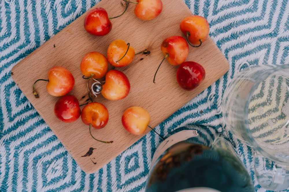 Fresh red cherries on wooden cutting board beside glass of blue beverage on geometric pattern fabric.