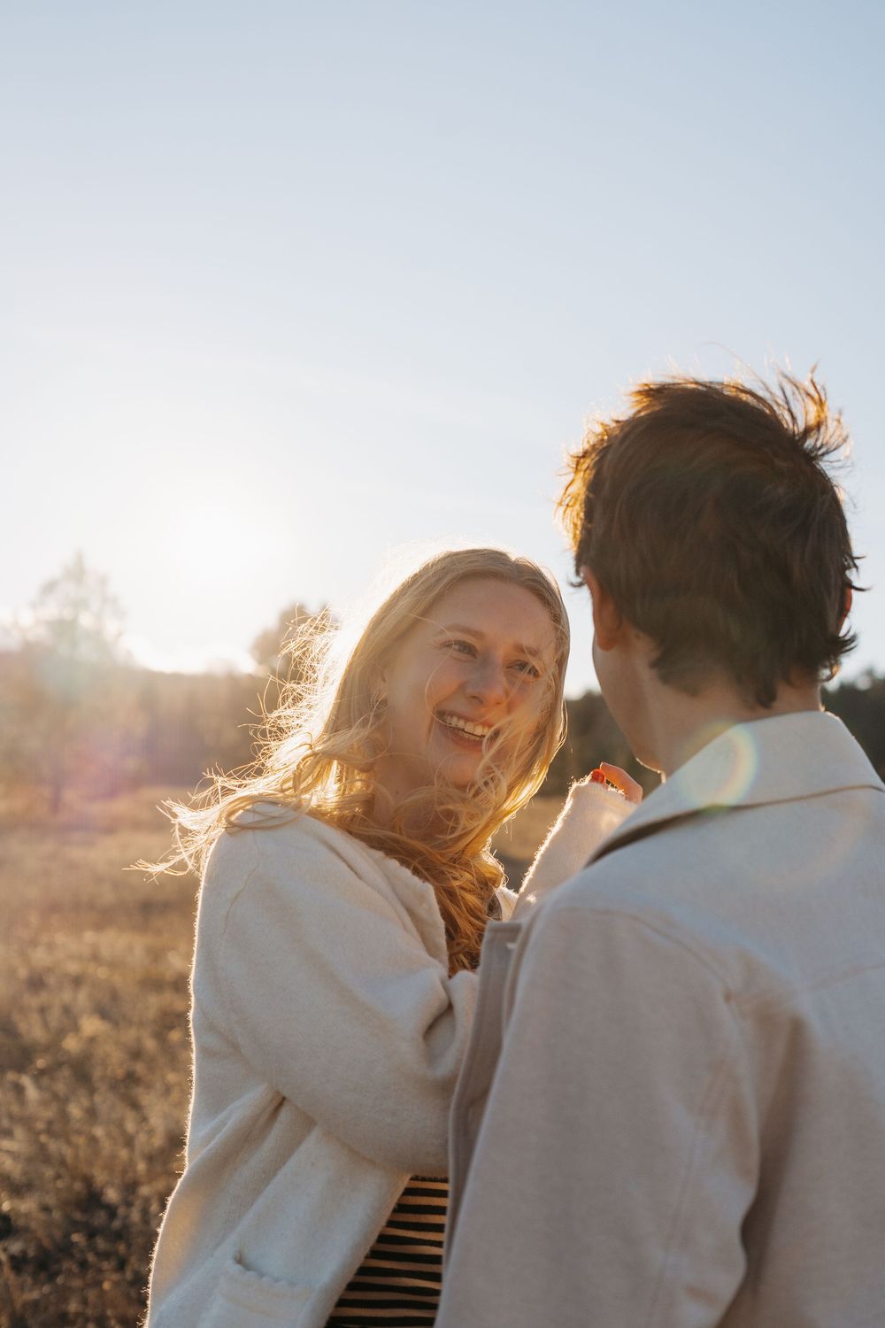 A smiling blonde woman and a man share a warm moment outdoors in golden sunlight during sunset.