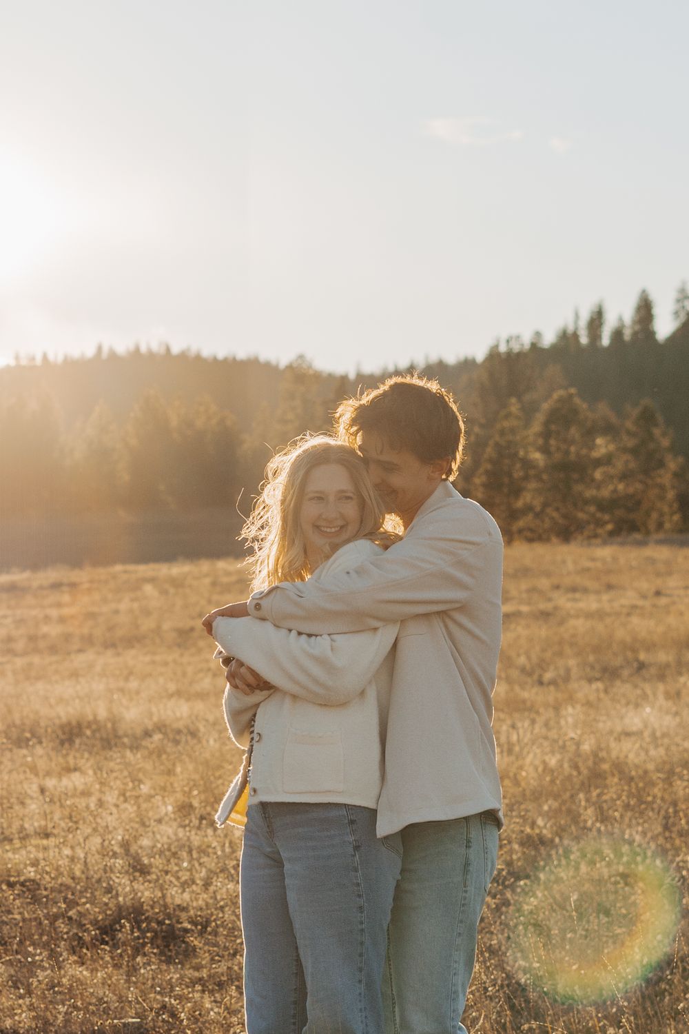 Couple embracing in golden sunlit meadow with forest backdrop during sunset, wearing casual beige and denim outfits.