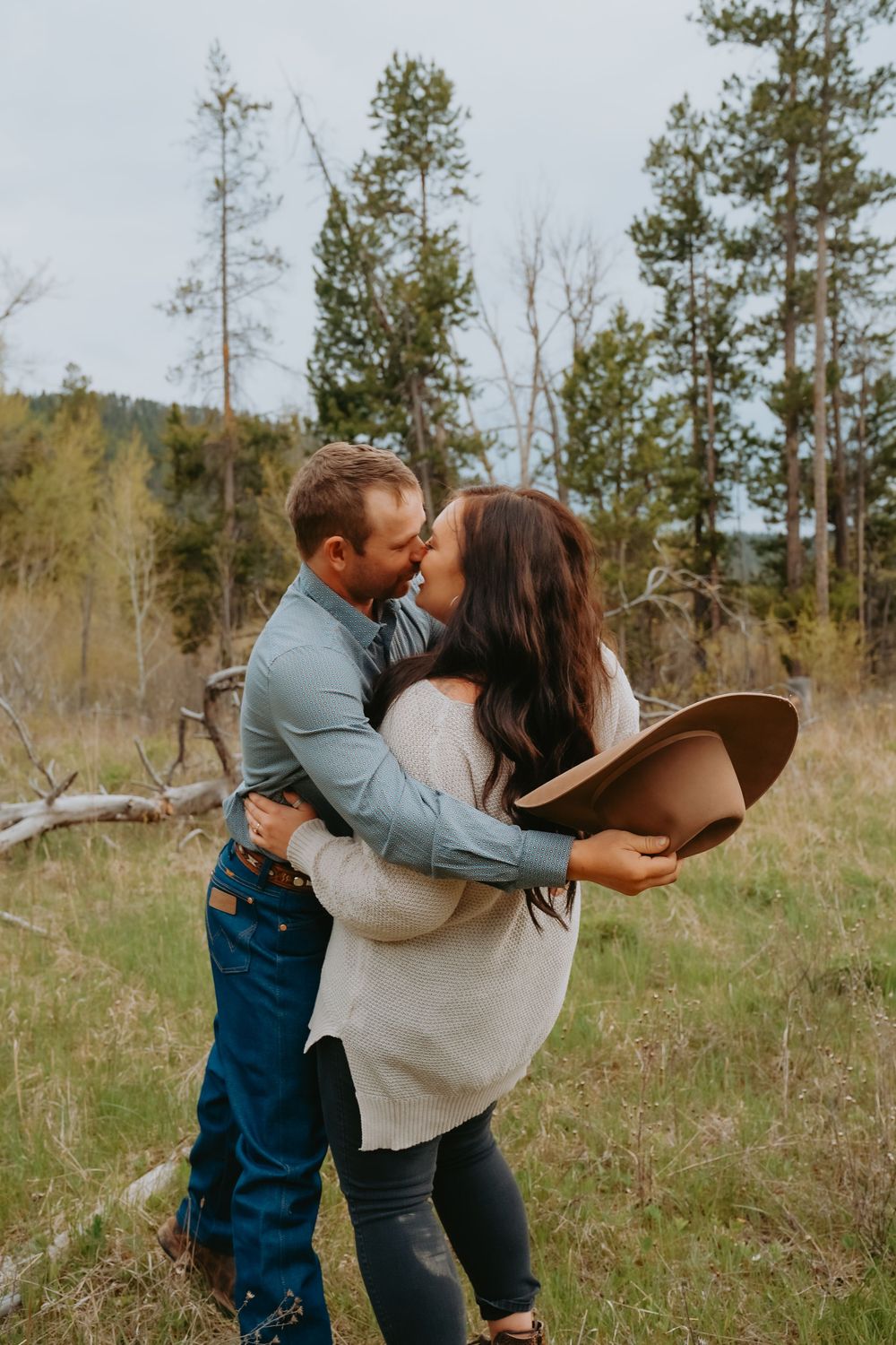 A romantic couple shares a tender moment in a wooded field while holding a brown hat.