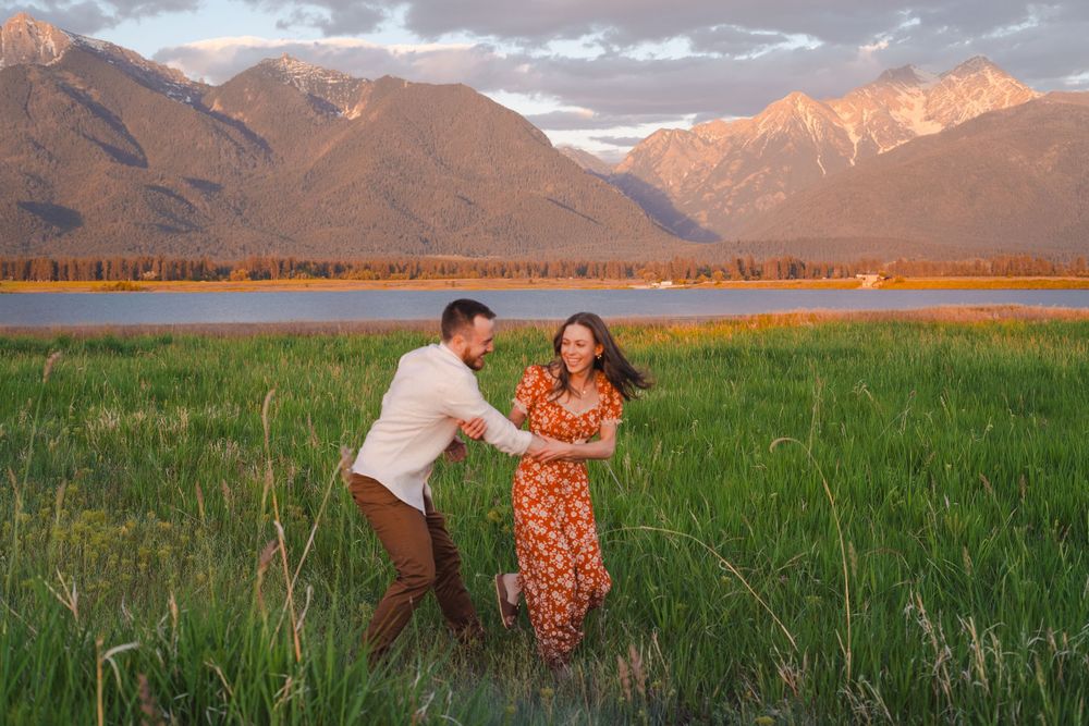 A couple dances playfully in a lush green meadow with majestic mountains and sunset-lit clouds in the background.