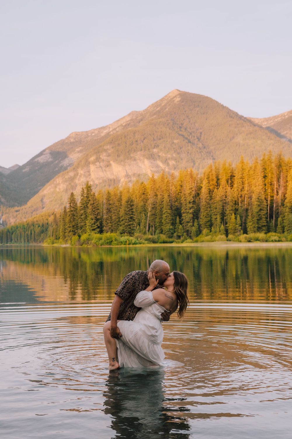 Two figures embrace while standing in a mountain lake with evergreen forests and peaks at sunset in Jasper National Park.