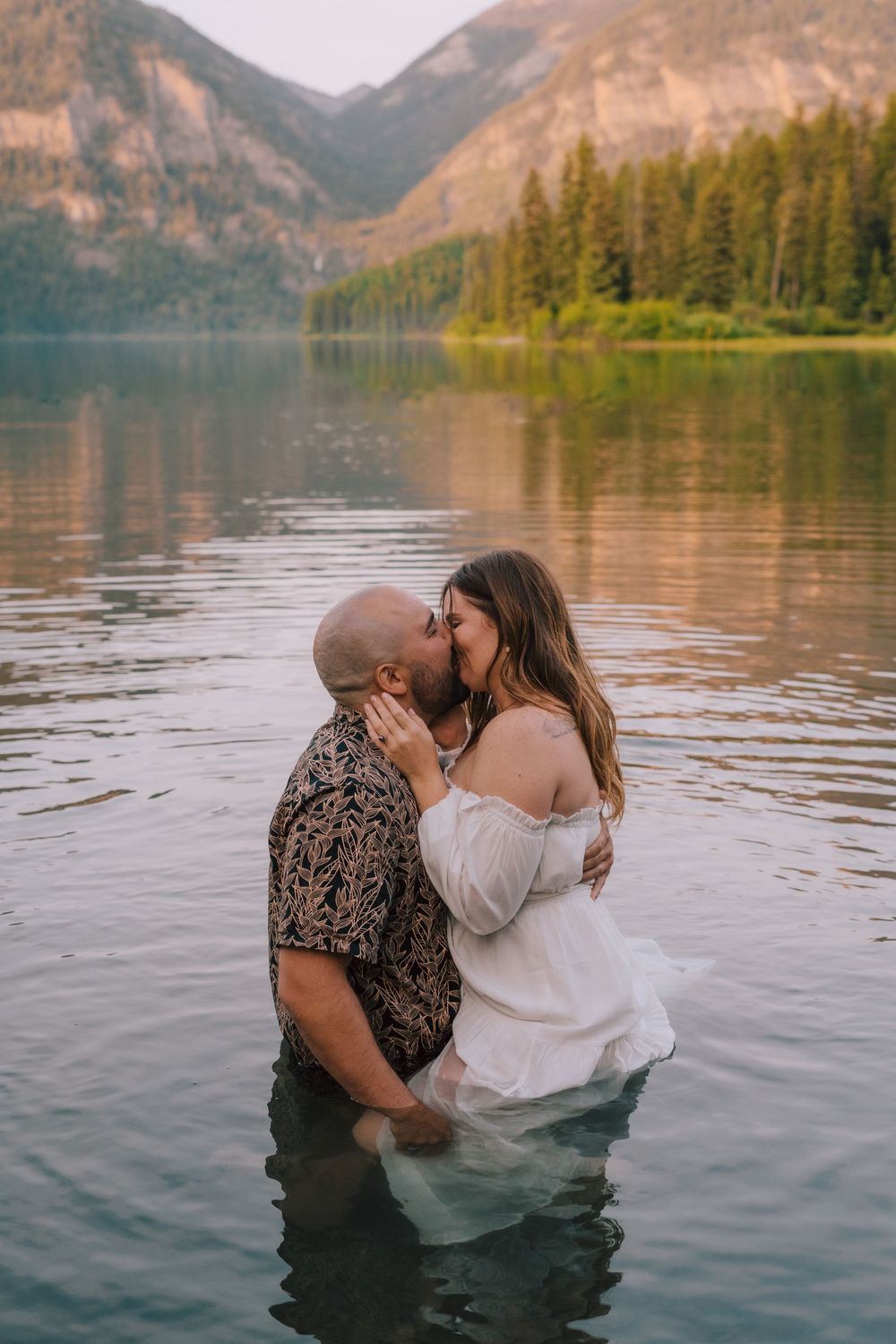 A romantic couple sharing intimate moments in a scenic mountain lake with forest and peaks in the background.