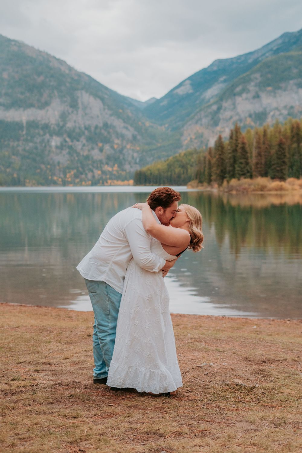 A romantic couple embraces by a serene mountain lake with towering peaks and evergreen trees in the background.