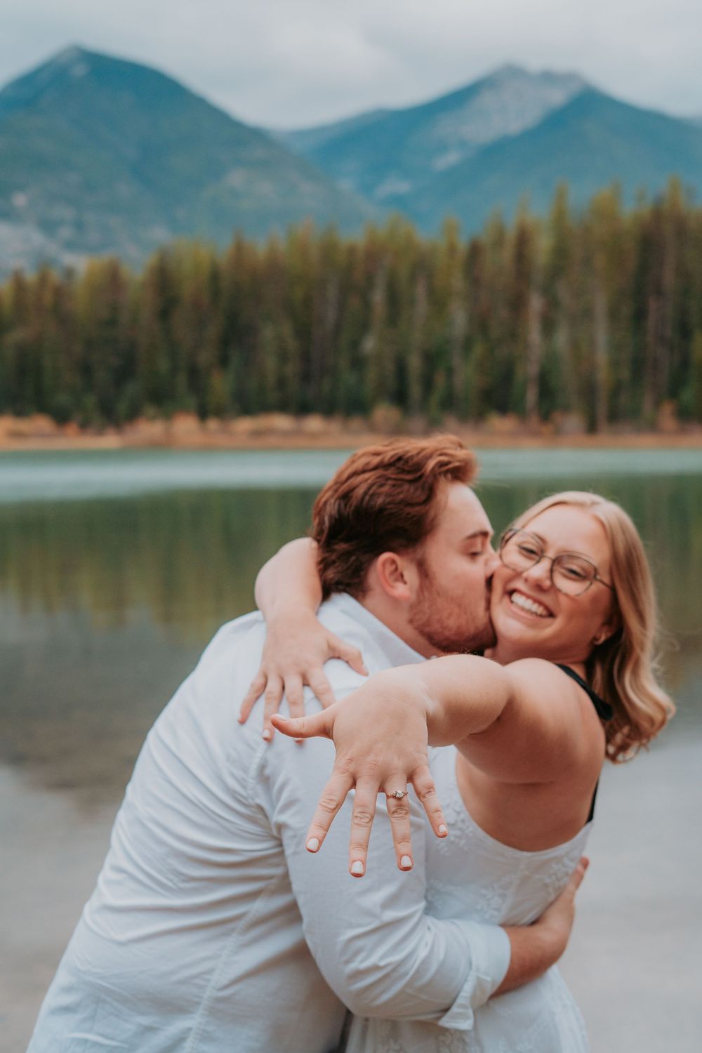 A romantic couple embraces and laughs together by a scenic mountain lake with evergreen trees in the background.