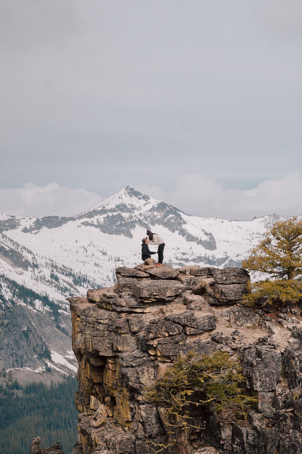 A person sits on a rocky cliff edge overlooking snowy mountain peaks and forested valleys in winter landscape.