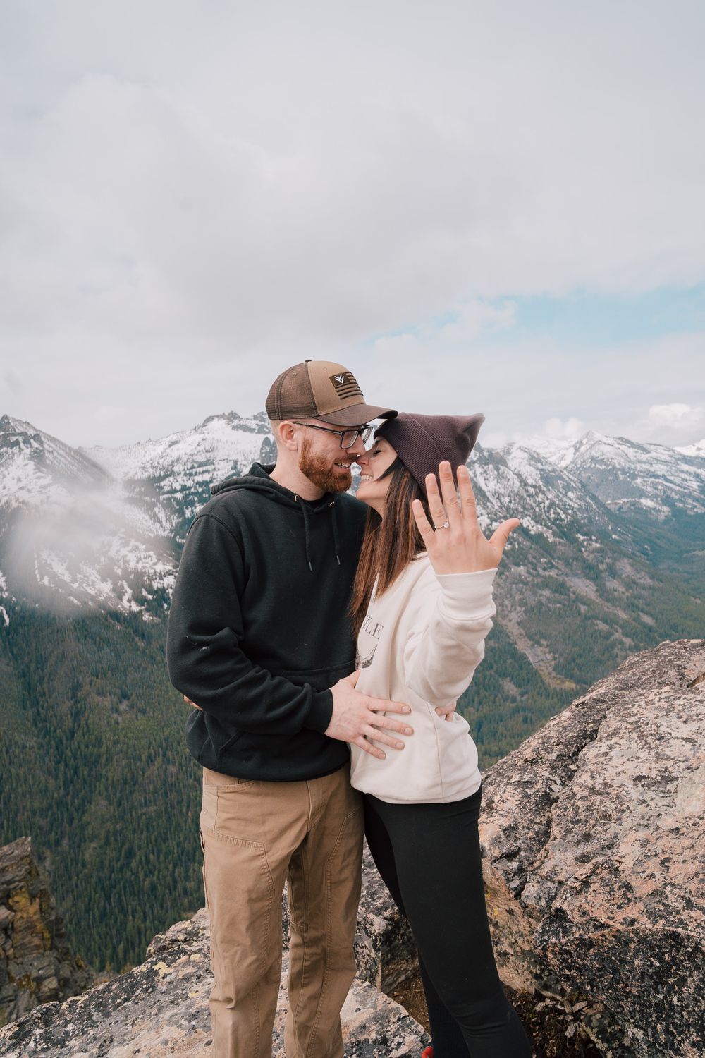 A couple shares a romantic moment at the summit of a mountain with snow-capped peaks in the background.