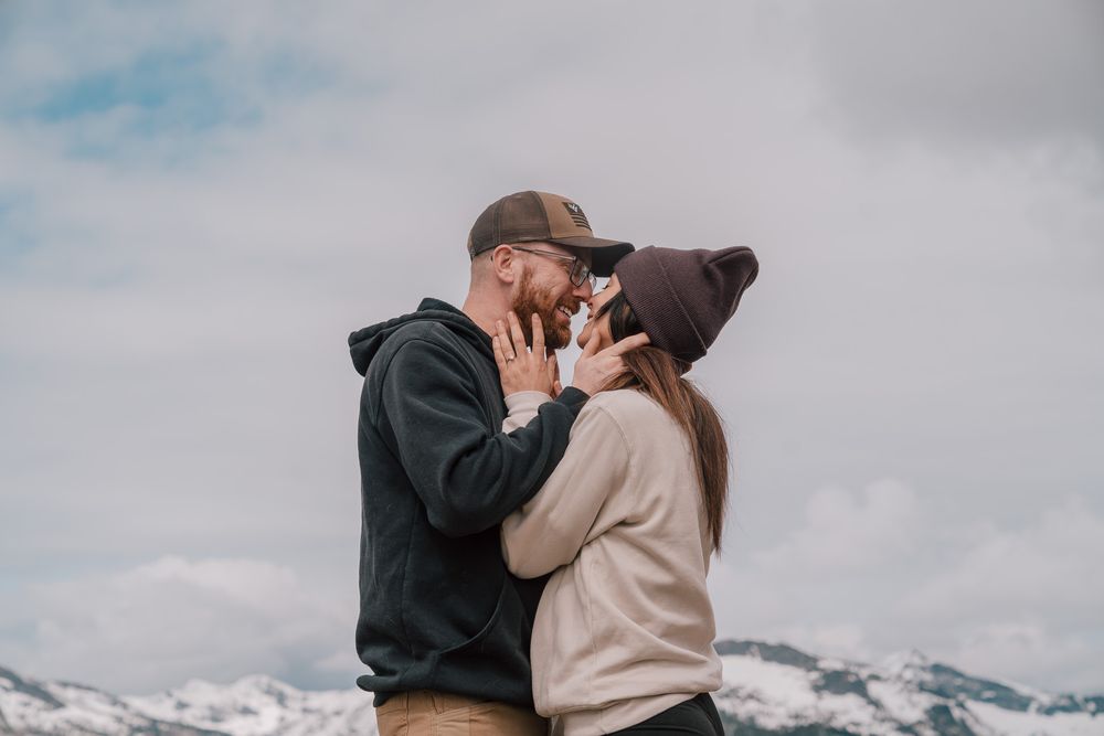 A couple embraces in front of a snowy mountain landscape while wearing casual winter attire on a cloudy day.