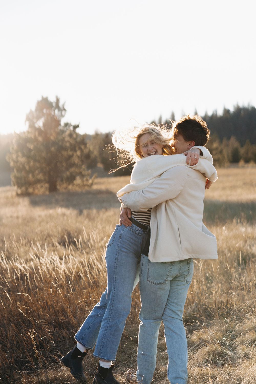 Couple embracing joyfully in a golden field at sunset, wearing jeans and white jackets, with trees in the background.
