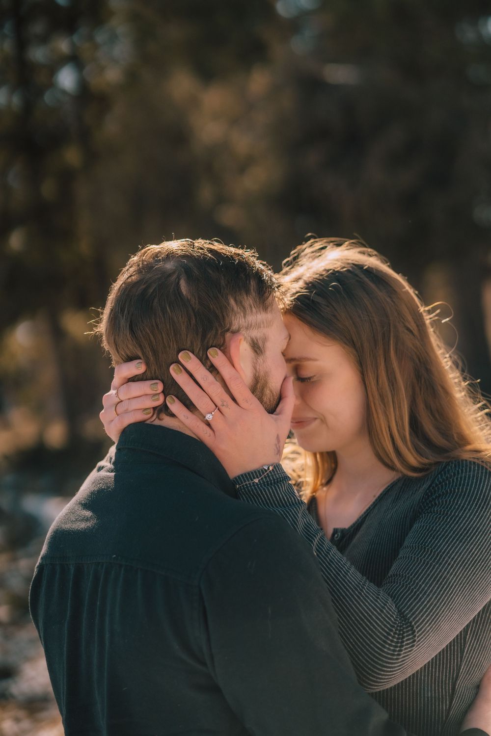 Romantic couple embracing outdoors, woman with engagement ring gently holding man's face in warm golden sunlight.