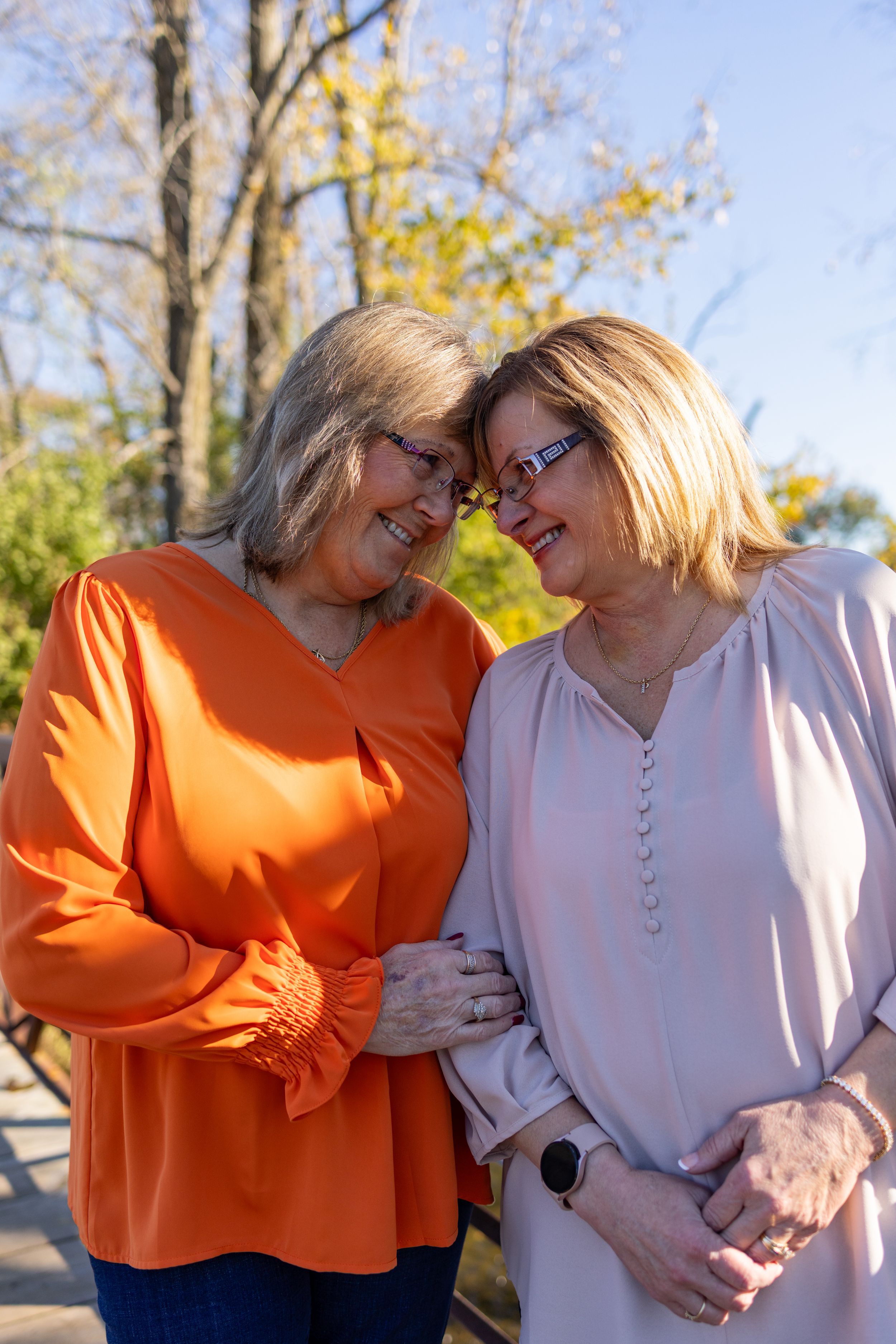 Two smiling middle-aged women in orange and pink blouses lean heads together outdoors in autumn setting.