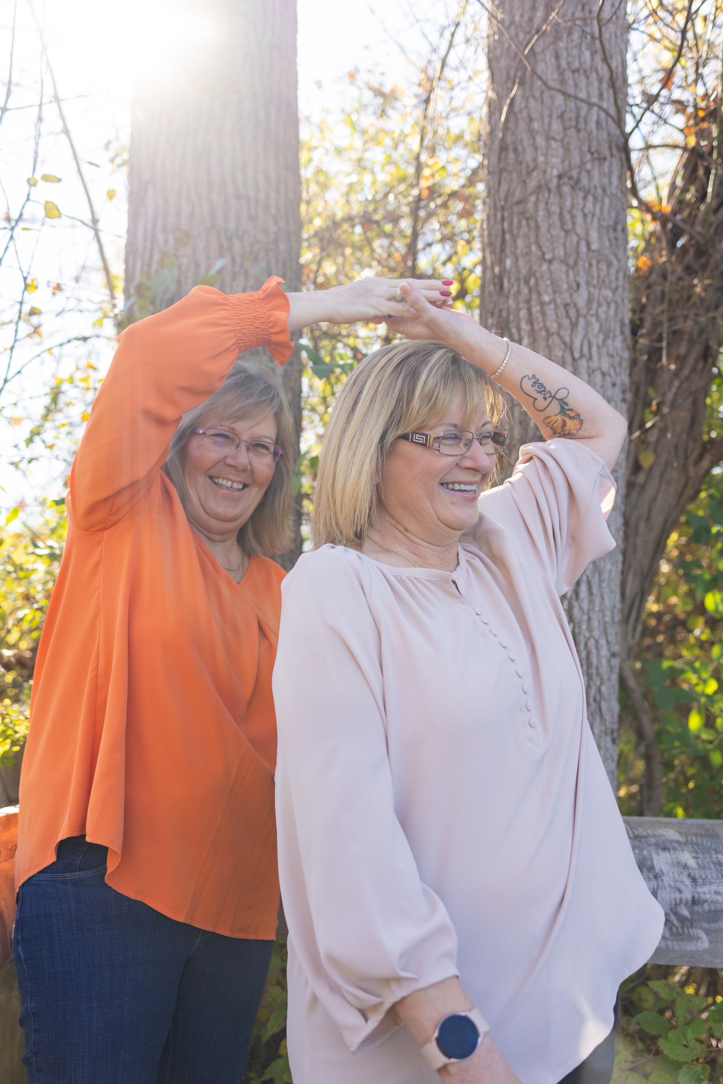 Two smiling women raise their arms outdoors among autumn trees in bright sunlight, one in orange and one in pink blouse.