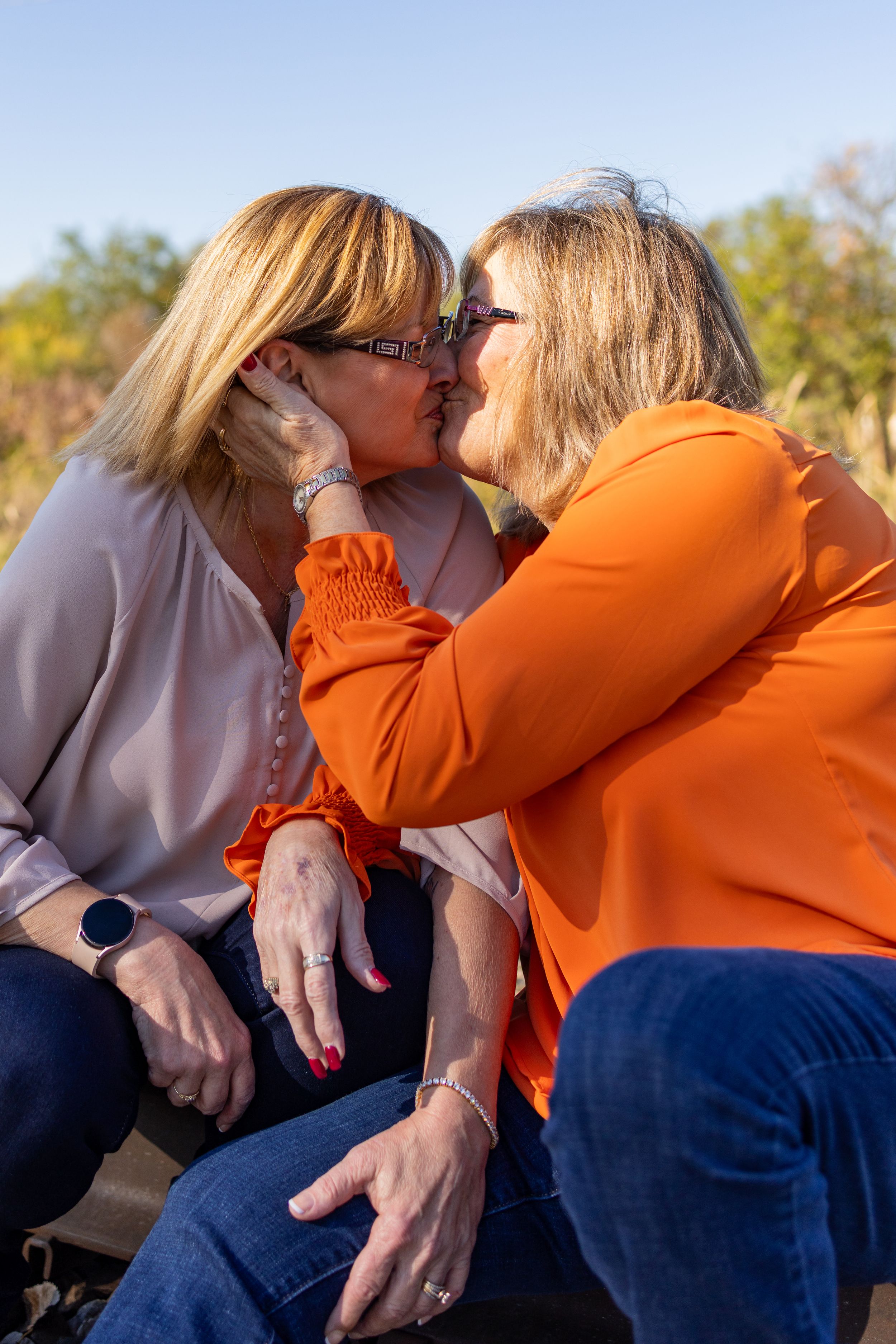 Two mature women sharing a kiss while seated outdoors on railroad tracks on a sunny day.