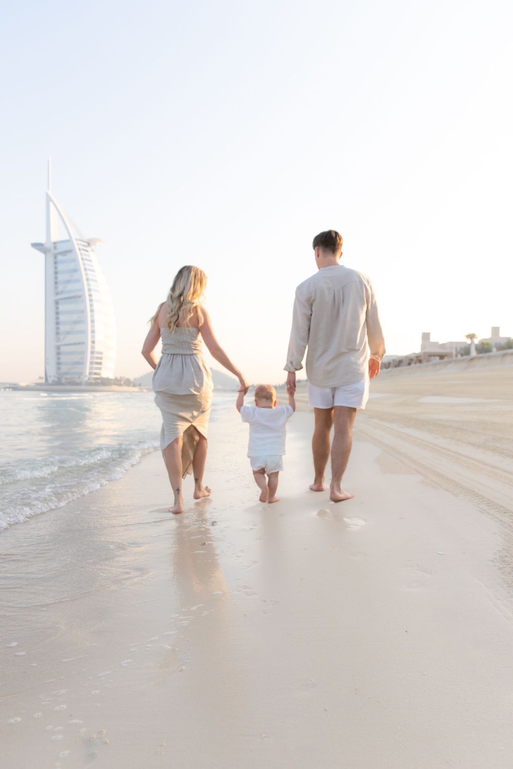 Parents with young toddler walking along the beach at sunrise with the Burj Al Arab in the background during outdoor family photoshoot