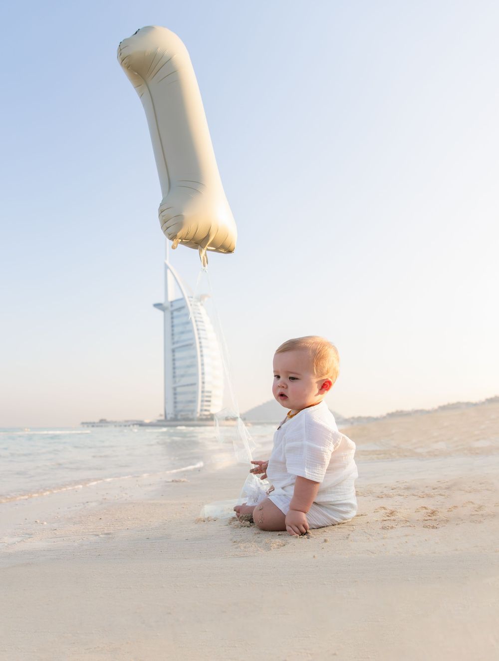 Baby sits on the beach with a 1st birthday balloon with the Burj Al Arab in the background during a sunrise outdoor photoshoot in Dubai