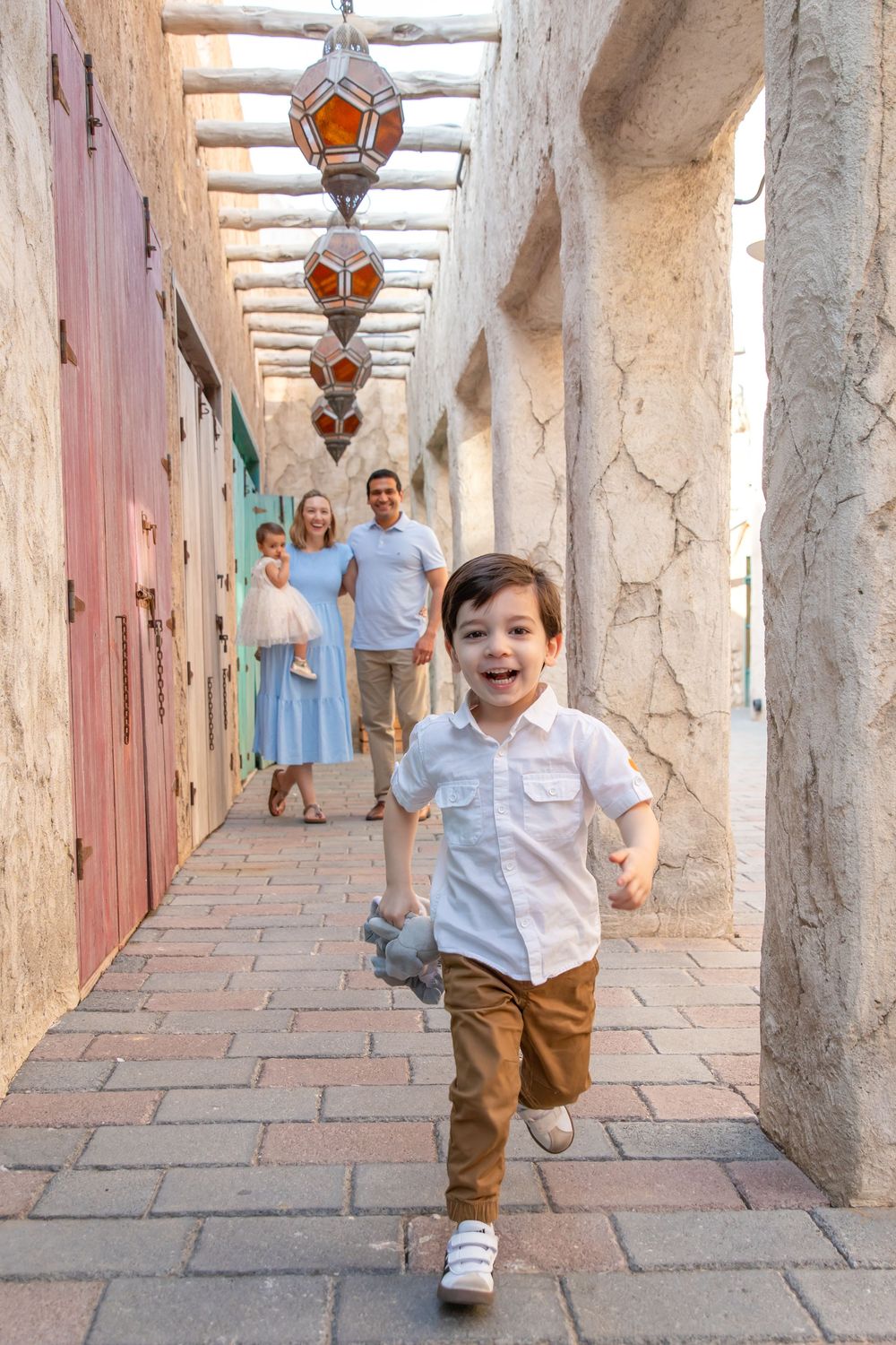 Young boy running down a colourful alleyway in Old Dubai with parents laughing in the background during family photoshoot in Old Dubai