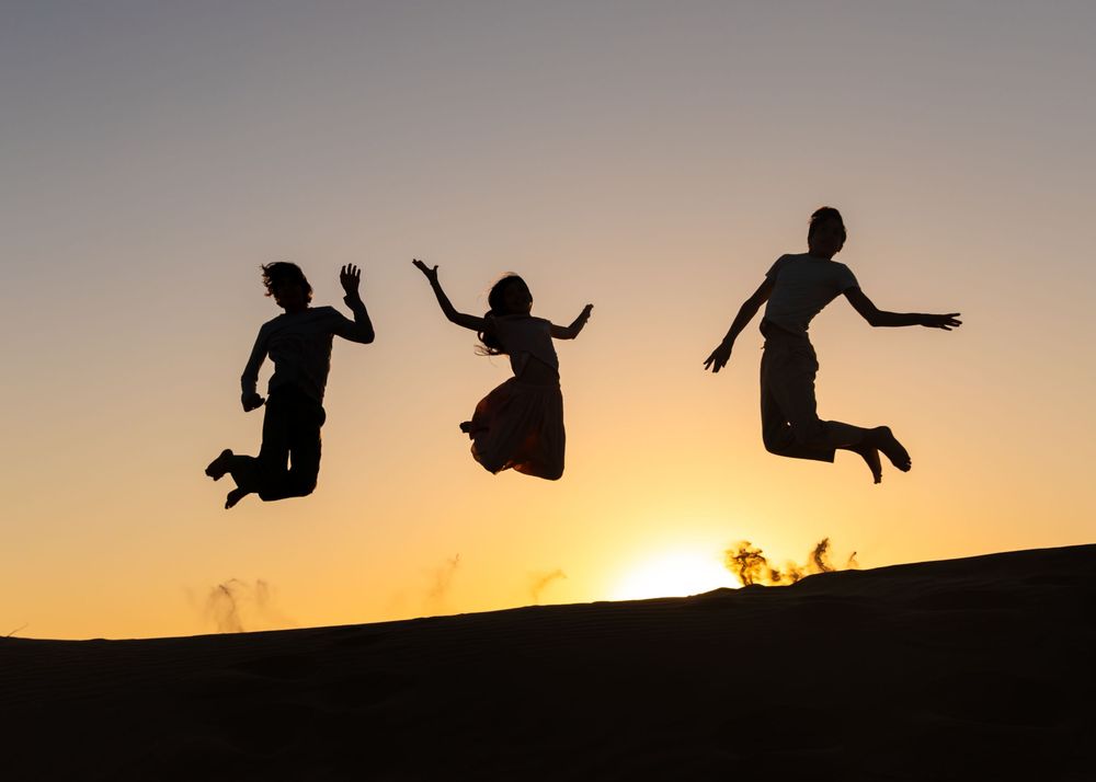Silhouette shot of 3 children jumping over the dunes with sunsetting in the background in Dubai desert during natural outdoor family shoot