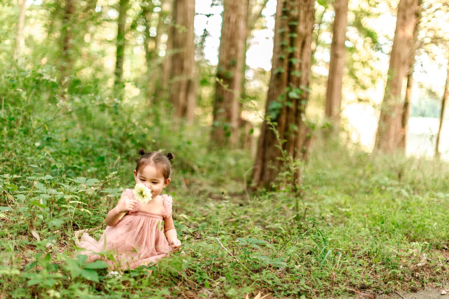 One-year-old girl in a pink tutu dress sniffs a flower in the middle of a whimsical woods setting.