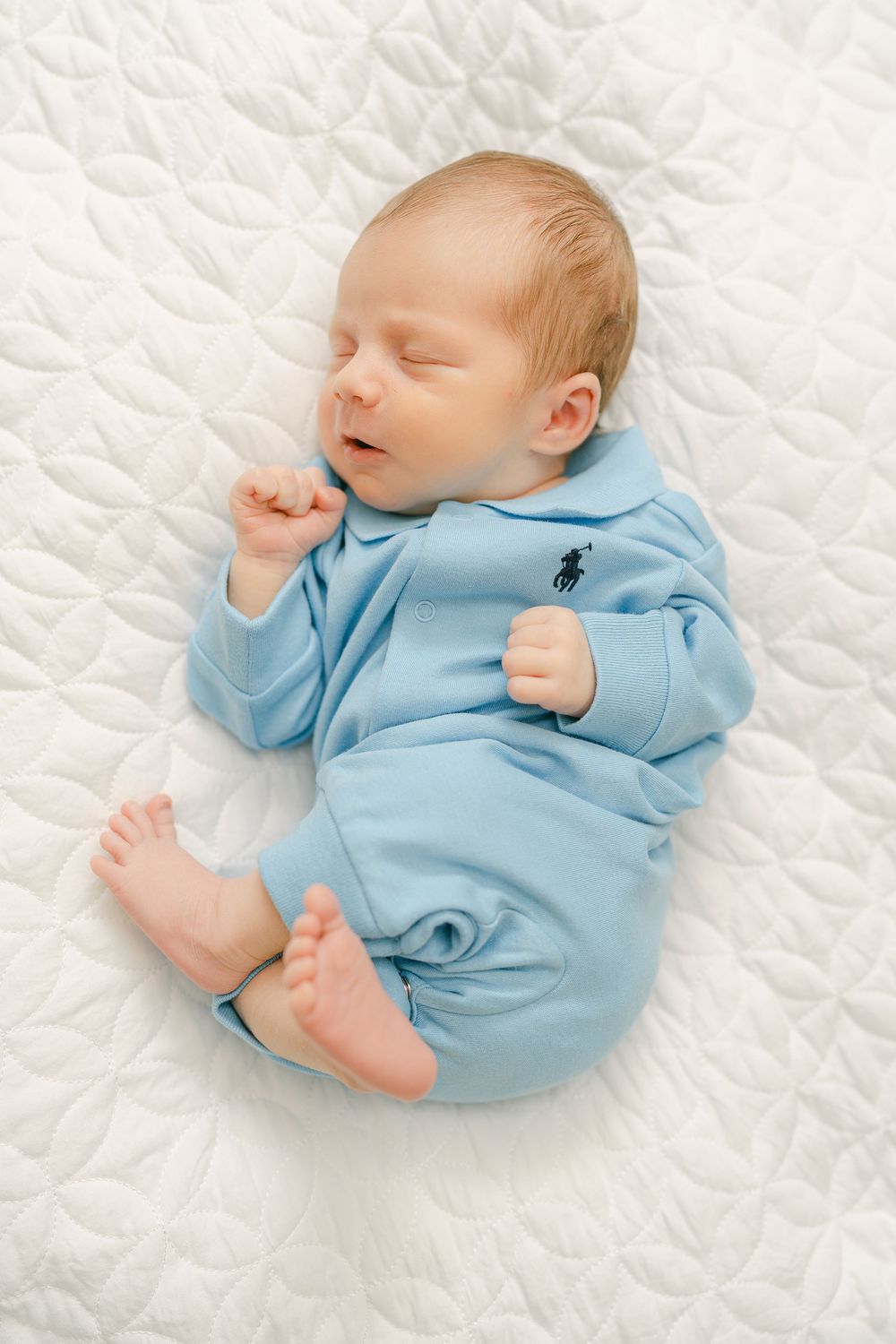 A sleeping newborn baby wearing a light blue onesie rests peacefully on white quilted bedding.