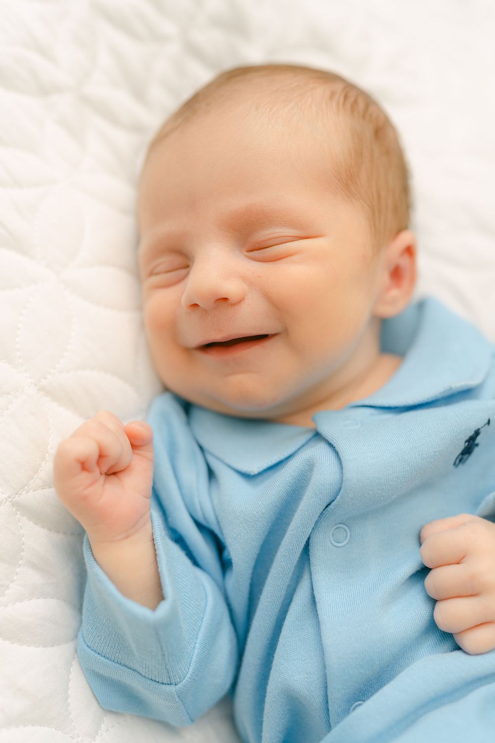 A sleeping newborn in a soft light blue onesie smiles happily while resting on white bedding.