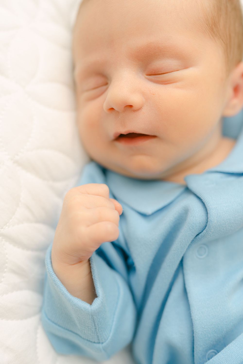 A sleeping baby wearing light blue clothing smiles contentedly while lying on white bedding.
