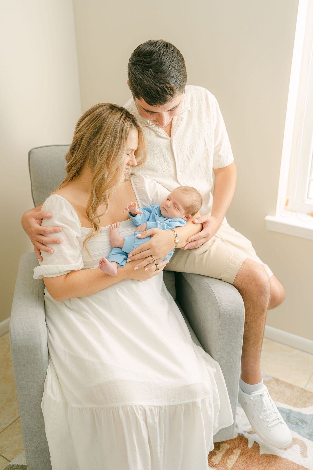 A tender family moment is shared on a cozy grey couch as they gaze lovingly at their newborn baby in soft natural lighting.