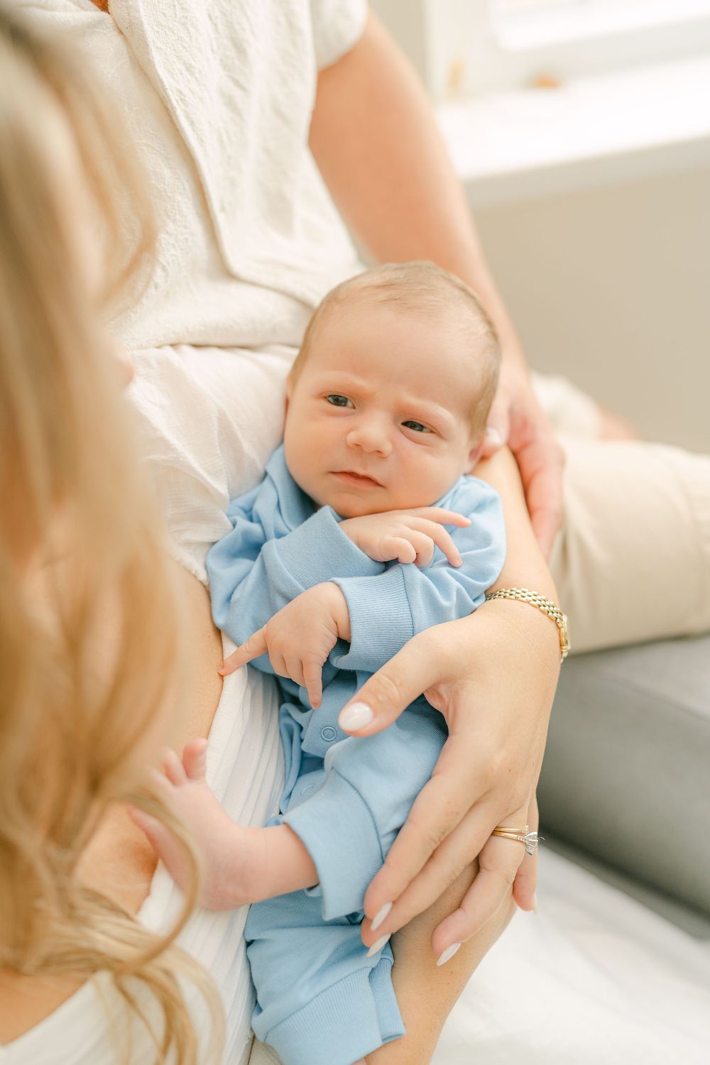 A newborn baby wearing a light blue outfit is gently held by caring hands in a soft, bright indoor setting.