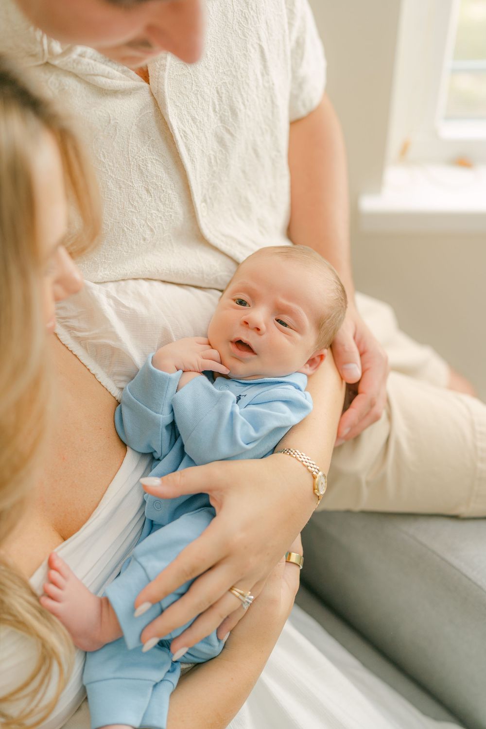 Baby wearing light blue onesie lies cradled in parent's arms near grey couch in sunlit room.