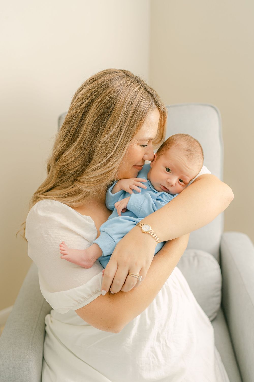A tender moment shows a mother embracing a sleeping baby in a white and blue ensemble on a light gray couch.
