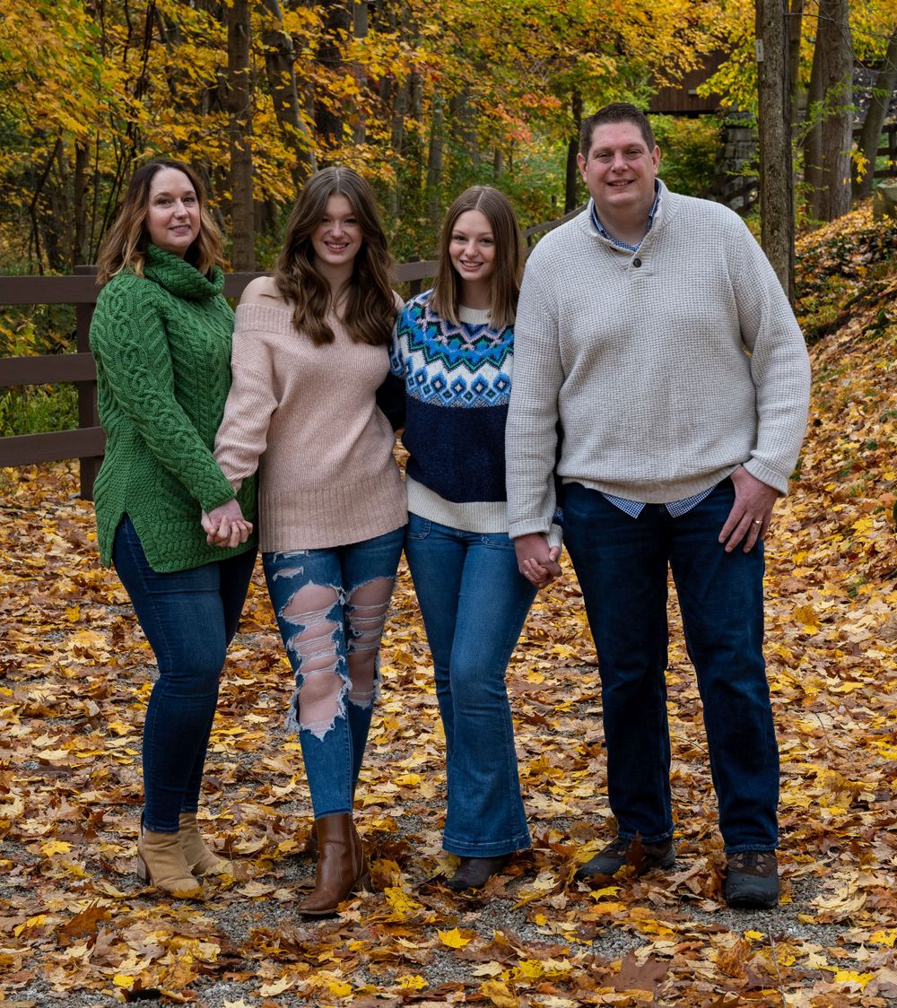 A family dressed in cozy sweaters poses together amidst fallen autumn leaves in a park setting.