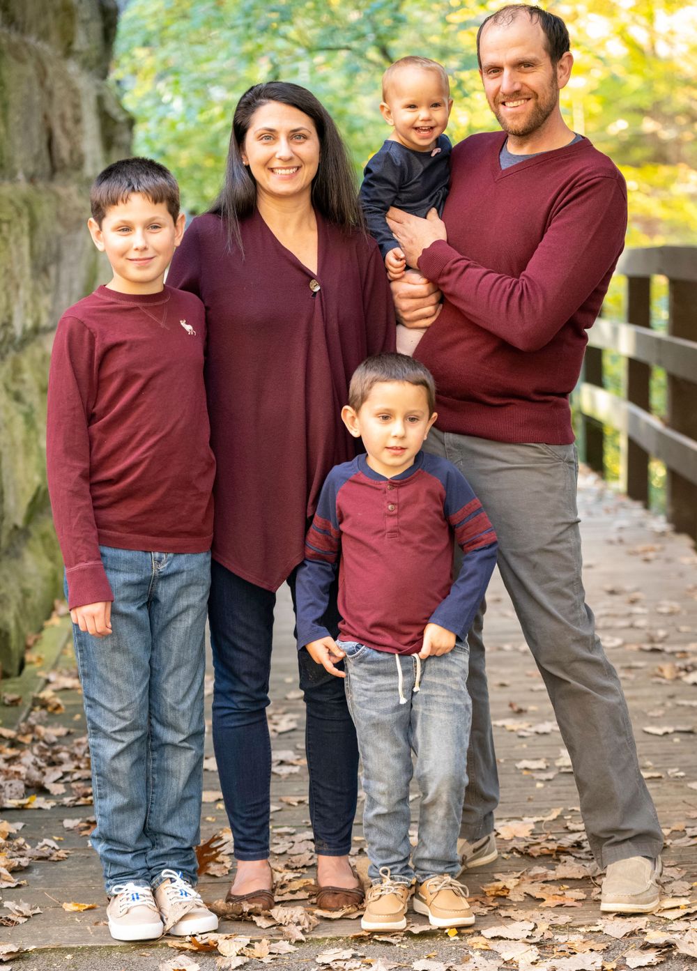 A family wearing coordinating burgundy sweaters poses on a wooden bridge during autumn.