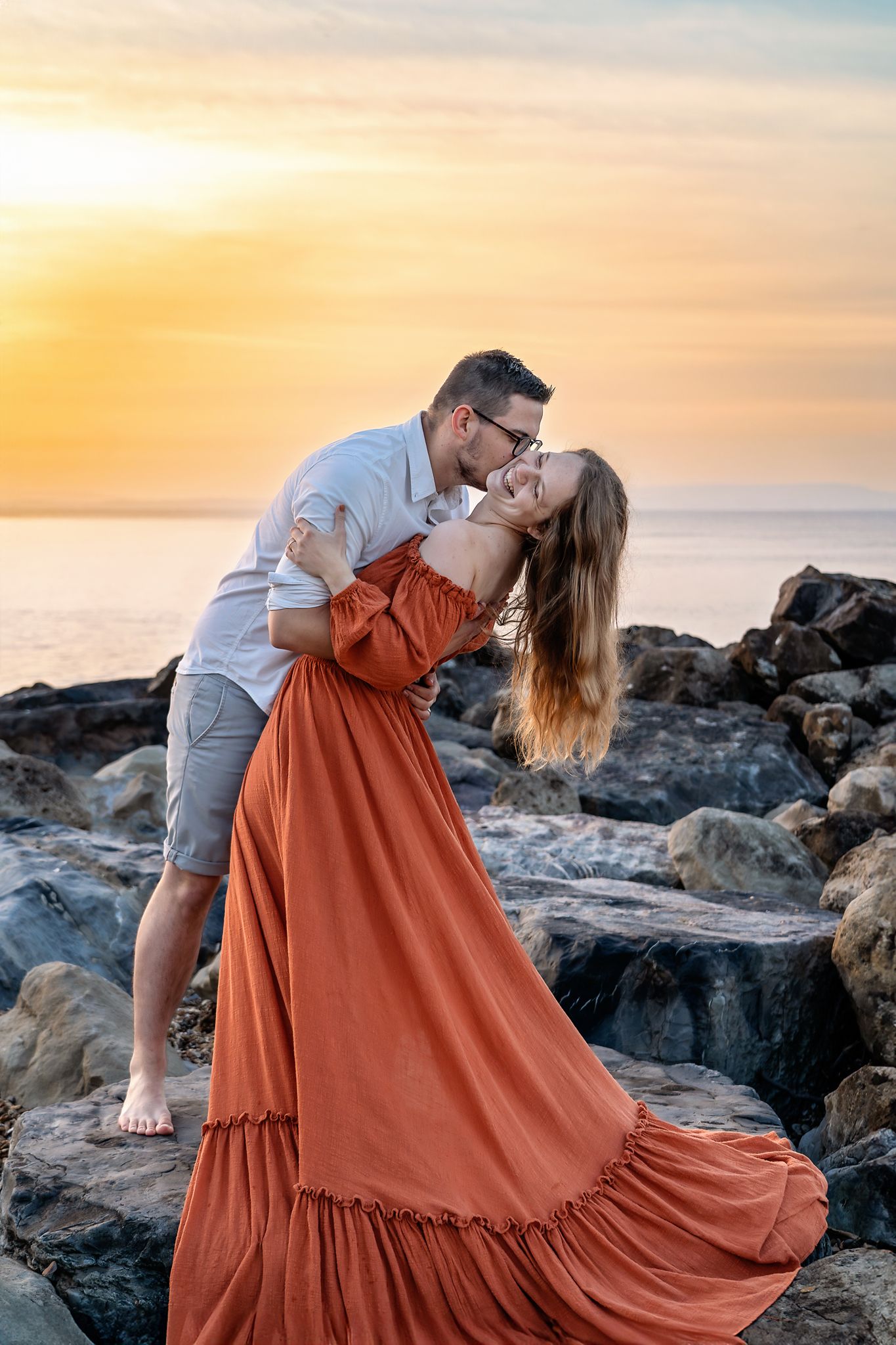 Barton on Sea engagement photoshoot. Couple hugging and kissing on rocks at sunrise, New Forest, UK.