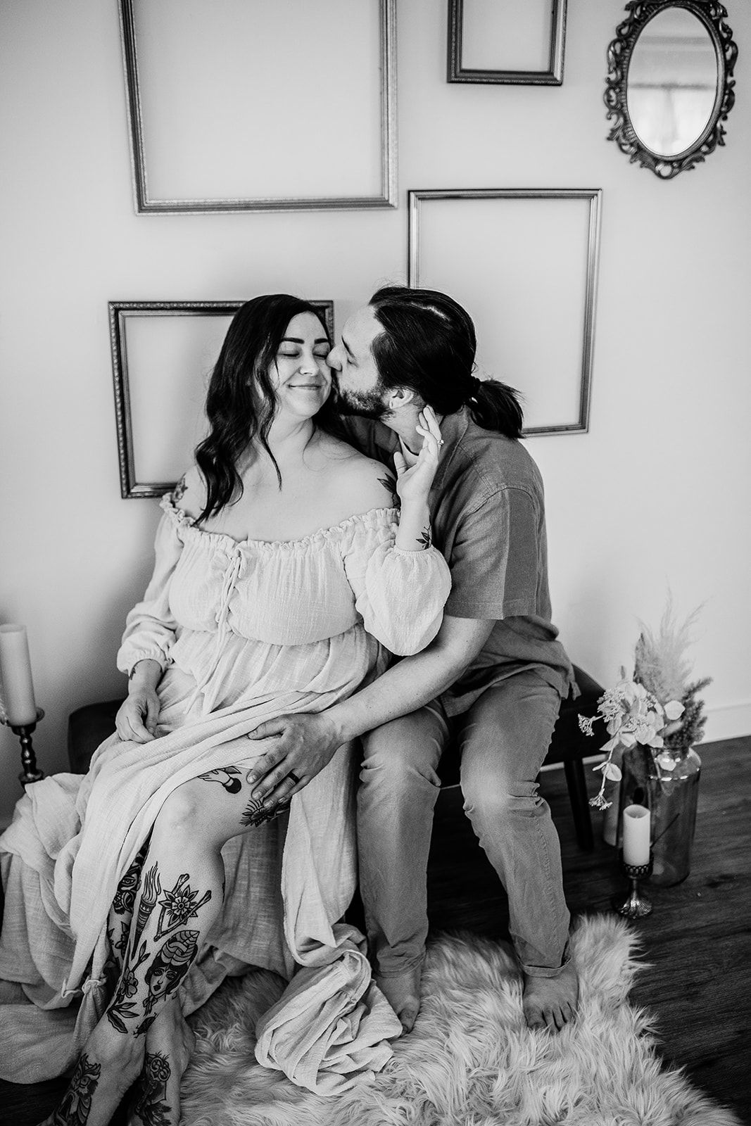 A man kisses a woman's cheek while she smiles, both seated closely together in front of empty picture frames on a wall.