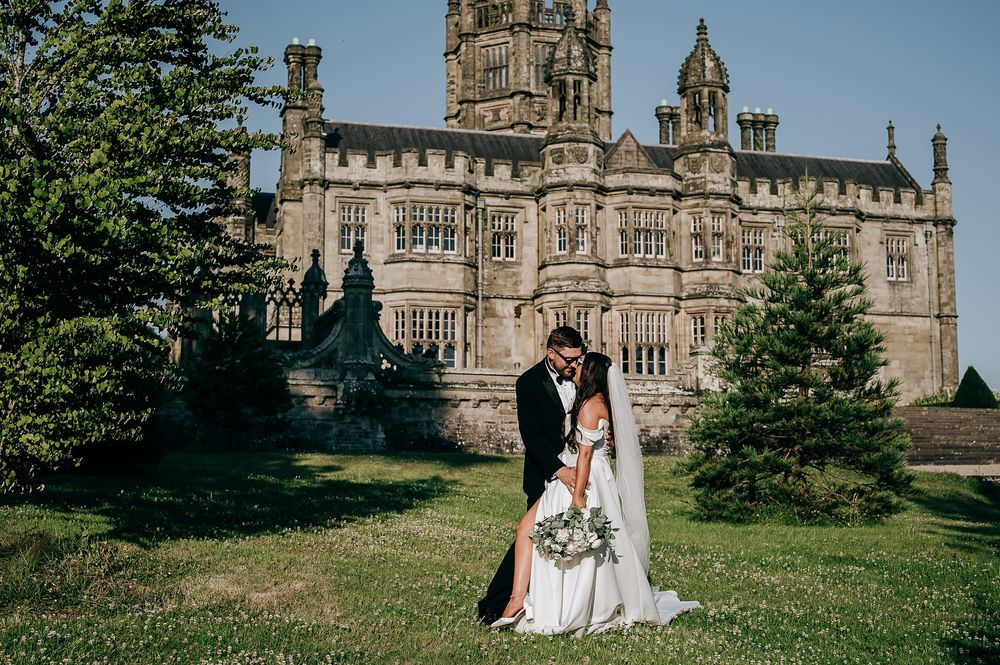 Bride and groom embrace on green lawn before grand historic stone castle on sunny wedding day.