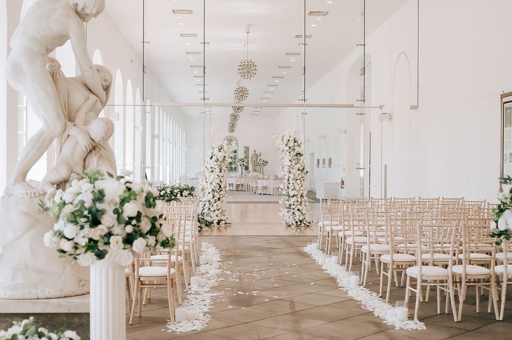 Elegant white wedding ceremony venue with floral arches, chiavari chairs, petal-lined aisle, and classical sculpture.