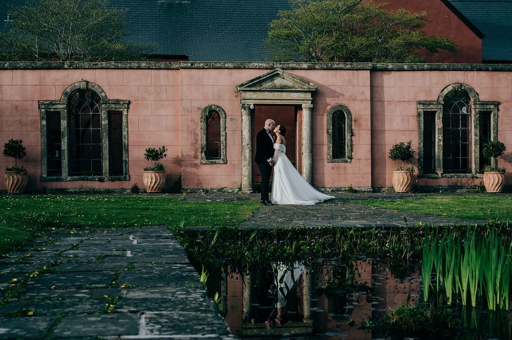 A couple stands before a pink stone building with arched windows reflected in a lily pond on their wedding day.