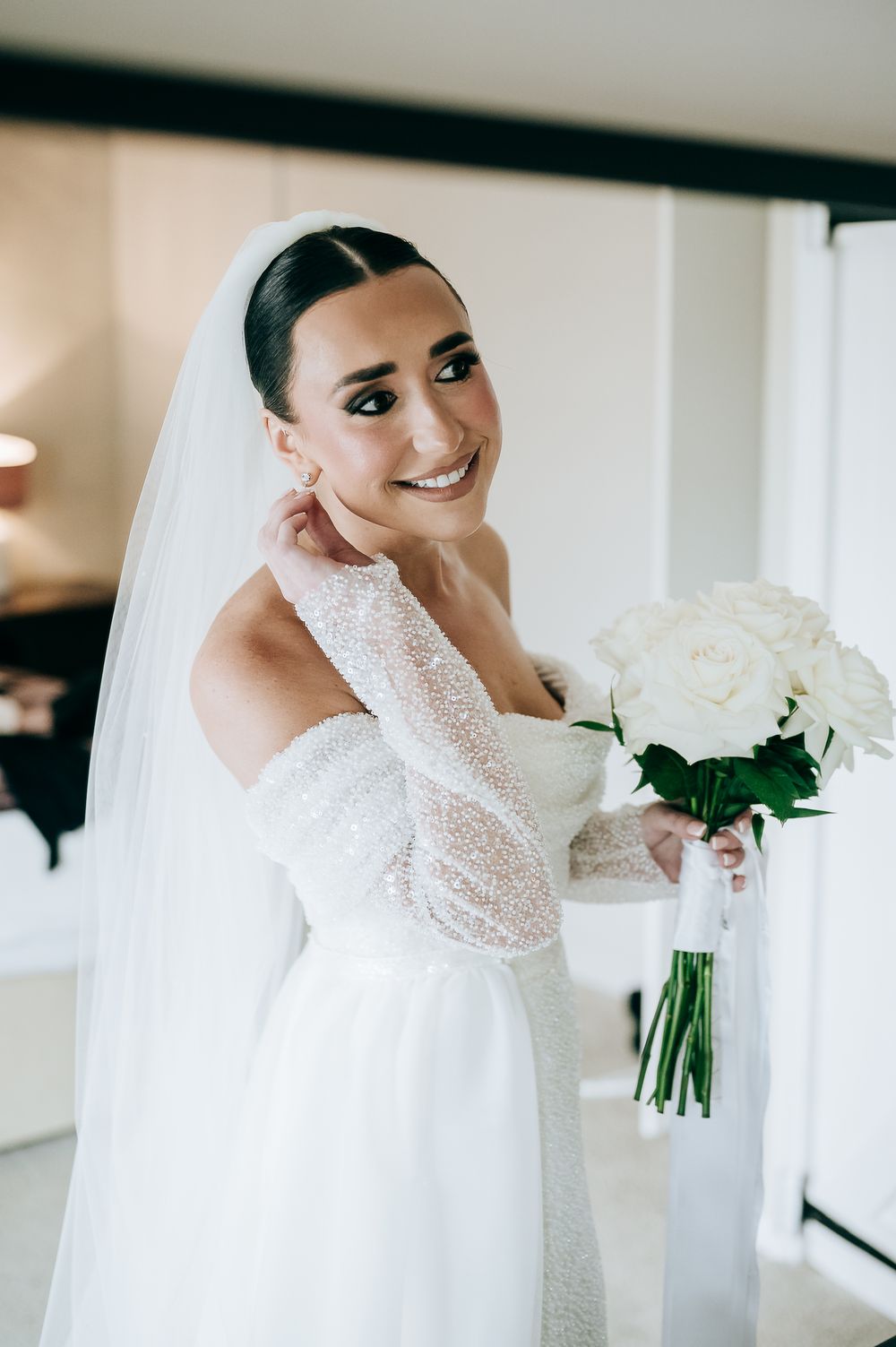 Elegant bride in lace off-shoulder gown holding white rose bouquet, wearing veil, smiling softly indoors.