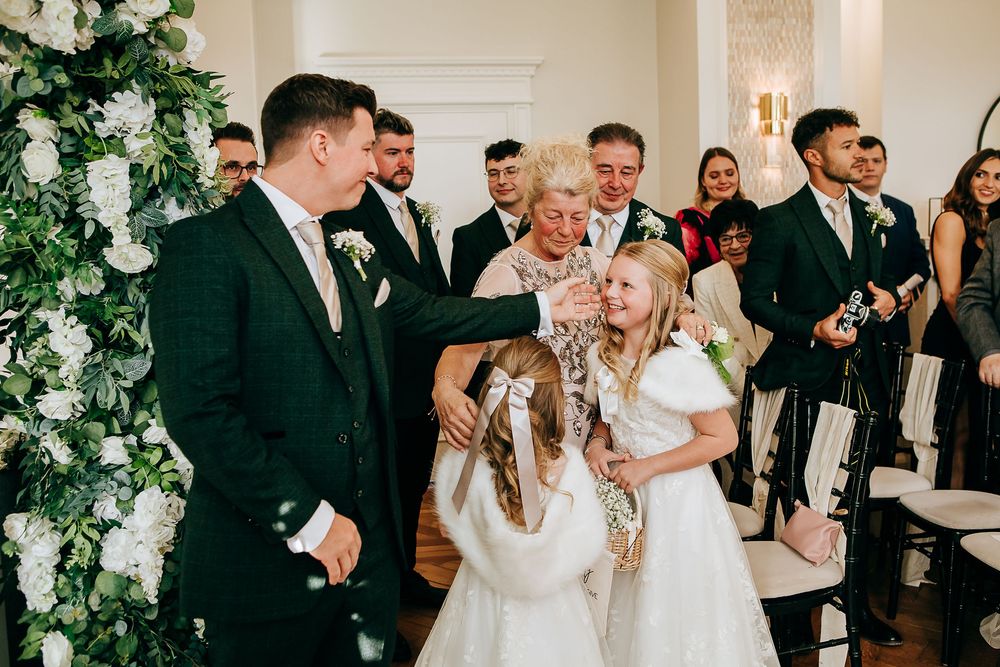 Groom in dark suit greeting flower girls during wedding ceremony with guests and floral arch in background.