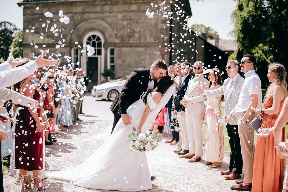 Bride and groom share romantic dip kiss while wedding guests toss white confetti outside historic church venue.