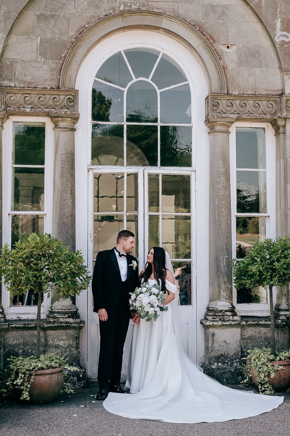 Bride and groom pose before an elegant arched window at a grand stone wedding venue, surrounded by lush potted topiaries.