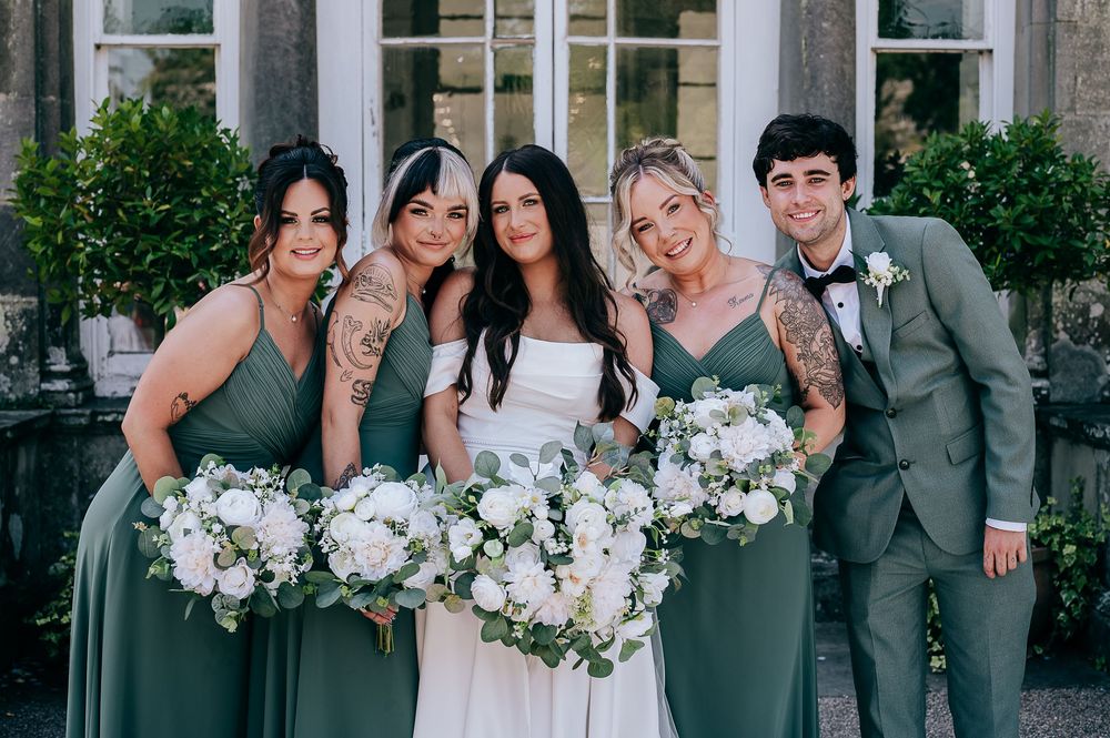 Bride in white gown with bridesmaids in sage green dresses and groomsman holding white floral bouquets at wedding.