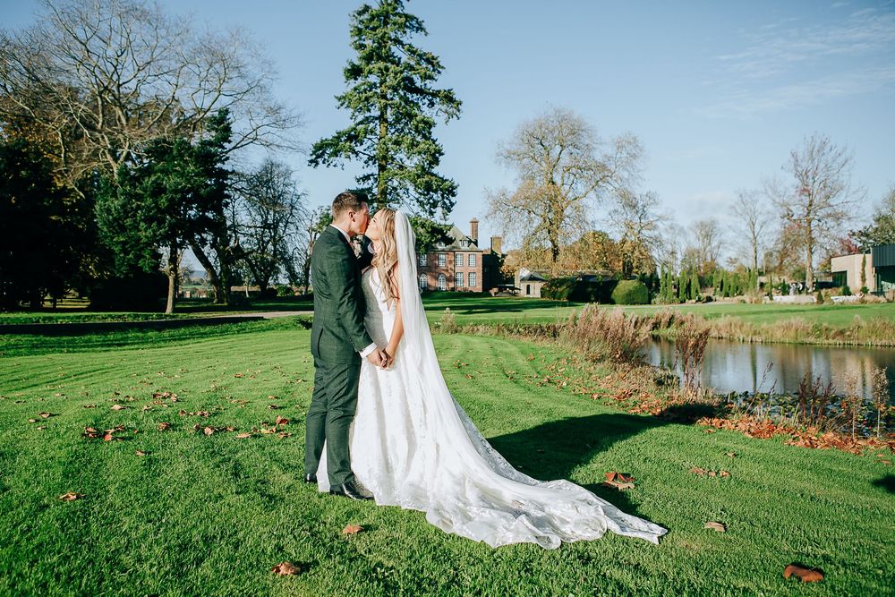 Bride and groom share a romantic kiss on lush green lawn beside a pond at an elegant countryside wedding venue.