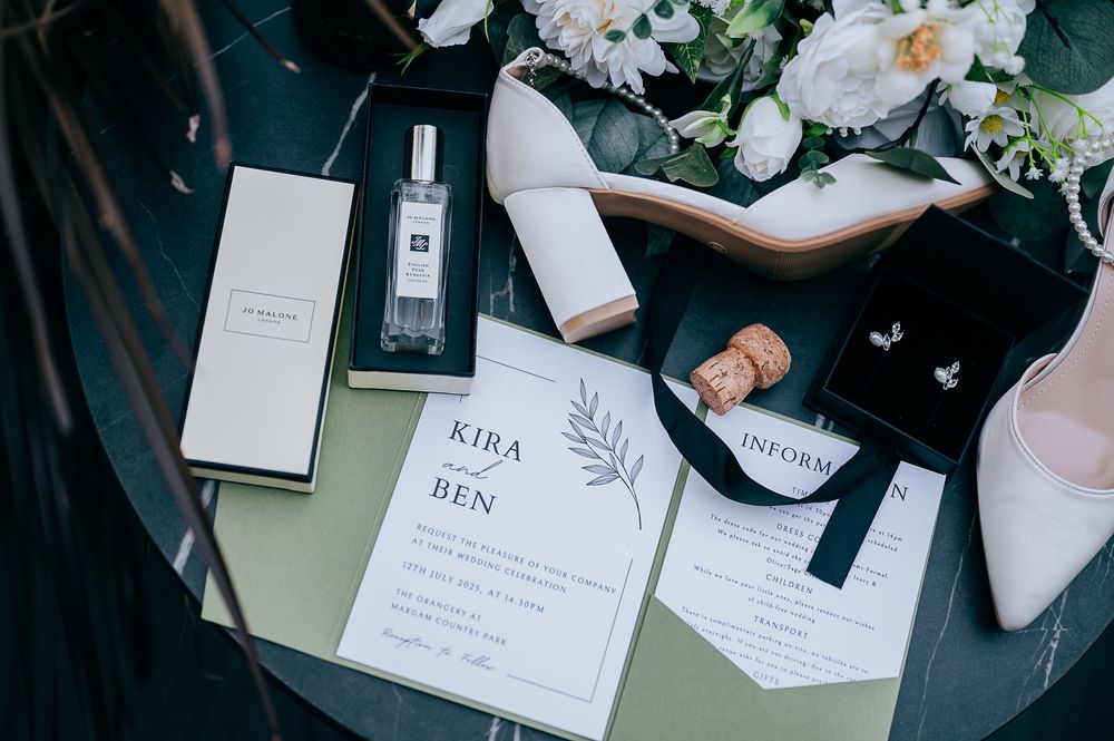Elegant wedding flat lay featuring Jo Malone perfume, white heels, floral bouquet, invitation suite for Kira and Ben, and diamond earrings.