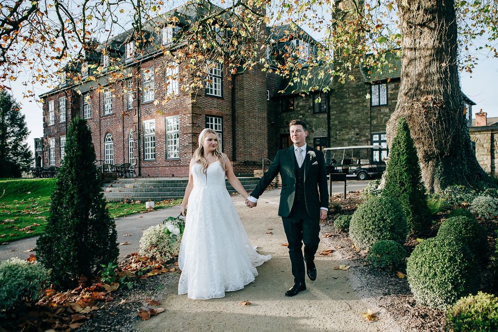 Bride and groom walk hand in hand outside a grand brick manor house surrounded by manicured hedges and autumn trees.