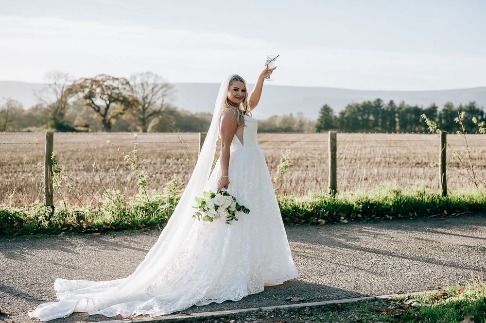 Bride in white lace gown with veil holding bouquet, posing on rural road with fields in background.