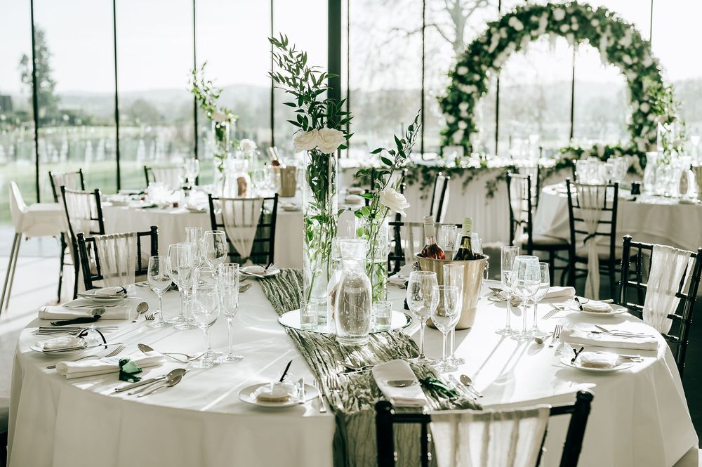 Elegant wedding reception with white tablecloths, crystal glassware, greenery centerpieces, and a floral arch in a bright venue.
