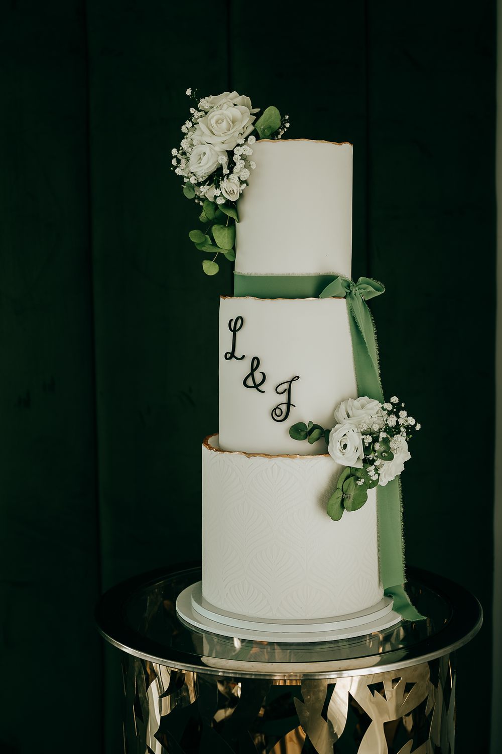 Three-tier white wedding cake with green ribbon, white roses, and L&G monogram on a gold stand.
