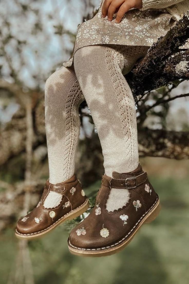 Girl sitting on tree branch wearing brown leather floral T-bar shoes with cream knit tights.