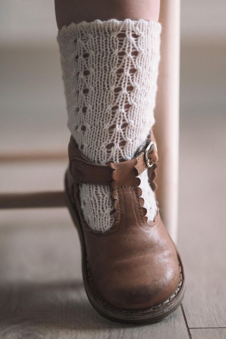Child's leg wearing white lace knit boot cuff sock with brown leather T-strap buckle shoe on wooden floor.