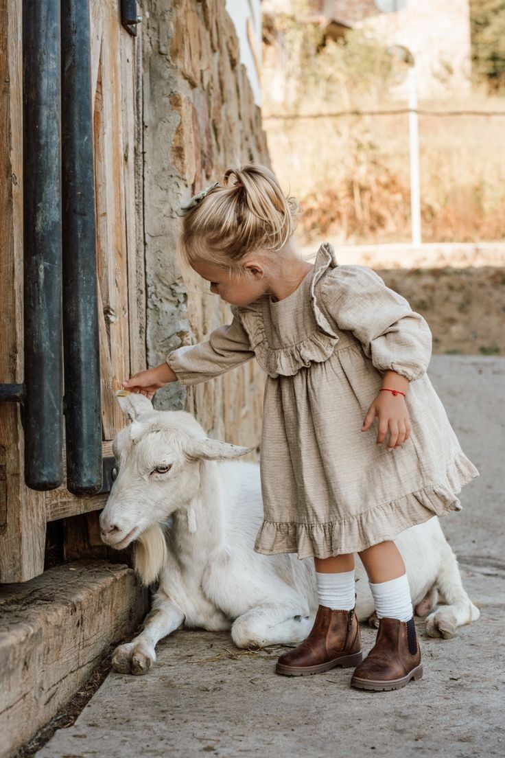 A young girl in a linen dress gently pets a white baby goat resting by a rustic wooden barn wall.
